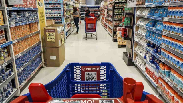 Persona comparando precios en su teléfono móvil frente a góndolas de un supermercado.